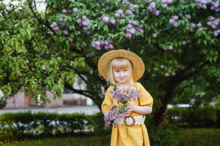A little girl in a yellow dress and straw hat wearing a bouquet of lilacs. A walk in a spring park, blossoming lilacs.の写真素材
