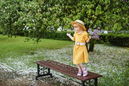 A little girl in a yellow dress and straw hat wearing a bouquet of lilacs. A walk in a spring park, blossoming lilacs.の写真素材