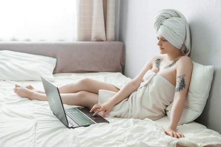 Tver, Russia-august 2, 2021 A woman after a shower is lying on the couch with a laptop and an eye mask at the same time. The concept of multitasking in the twenty-first century.のeditorial素材