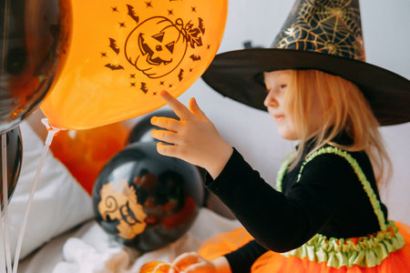 Childrens Halloween - a girl in a witch hat and a carnival costume with airy orange and black balloons at home. Ready to celebrate Halloweenの写真素材