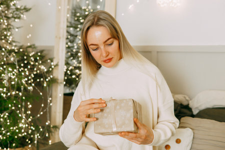 A blonde woman on a bed in a Scandinavian interior decorated for Christmas. A Christmas gift in a craft package. The period of the winter New Year holidays.の写真素材