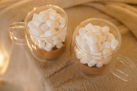 Two glass mugs of hot chocolate topped with marshmallows, placed on a soft beige blanket, with warm fairy lights in the backgroundの写真素材