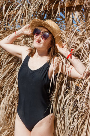 Woman on vacation near straw umbrella on beach. summer holiday concept. woman near palm leaves. person in a one piece swimsuit and sun hatの写真素材