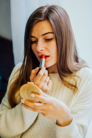 Woman applying red lipstick while looking in a compact mirror. Makeup application concept for beauty and cosmetic advertising.の写真素材