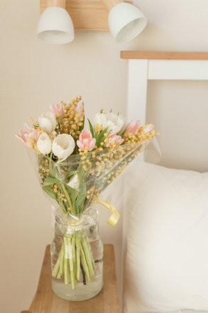 Bouquet of tulips and mimosa flowers in a vase on a bedside table. Interior decoration with fresh flowers in a home setting.の写真素材