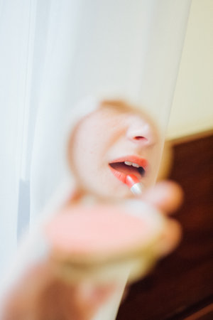 Woman applying vivid red lipstick using a compact mirror. Reflection of a female during morning makeup routine. Beauty procedure for cosmetic product.の写真素材
