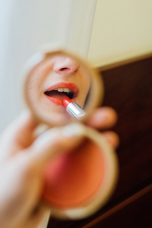 Woman applying vivid red lipstick using a compact mirror. Reflection of a female during morning makeup routine. Beauty procedure for cosmetic product.の写真素材