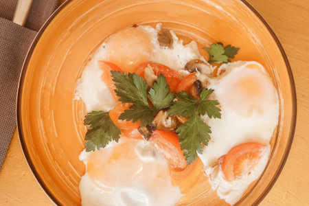 Close up view of fried eggs with tomatoes, mushrooms and parsley on a plate. Breakfast dish on a wooden table. Healthy and fresh morning meal for diningの写真素材