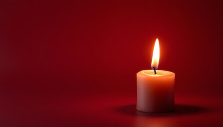 Single lit candle on a dark red background. Warm glowing flame providing light in a solemn or spiritual setting.の写真素材
