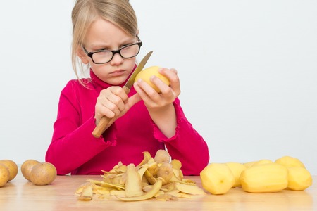 A girl is wearing a red turtleneck and glasses. It peels a potato with a sharp kitchen knife. Before him lies a heap of potato peelの写真素材