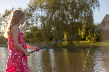 A 7 year old girl is standing by a lake with fishing rod and looking at the water. She waits for a fish to biteの写真素材