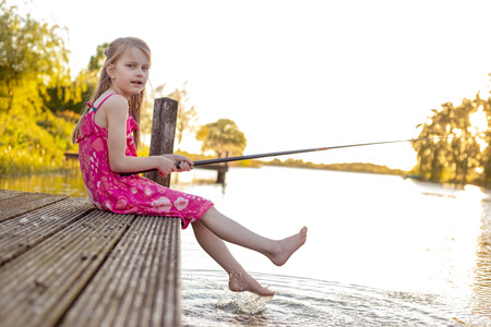 A 7 year old girl is sitting with her fishing rod on a jetty by a lake, waiting for a fish to bite. She looks into the camera and splashes with her feet in the waterの写真素材