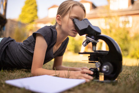 A 9 year old girl lies on the grass in the garden and looks into the eyepiece of a microscope. In front of her is a sheet of paper.の写真素材
