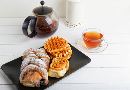 Black saucer with pastries and transparent cup of tea on white wooden table.の写真素材
