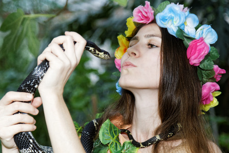 Young girl with floral wreath on her head holding a snake in his hands. Foliage forest on dark background.の写真素材