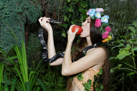 Young girl with floral wreath on her head biting an apple in one hand and holding a serpent in the otherの写真素材