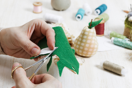 Handicrafts. Closeup hands of the needlewoman hold scissors and cut out a piece of pattern for a handmade toy. There are handicraft materials on wooden table.の写真素材