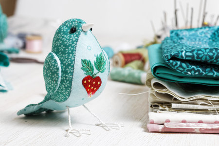 Closeup a handmade bird sewed from a cloth stands on a white wooden table surrounded by handicraft...