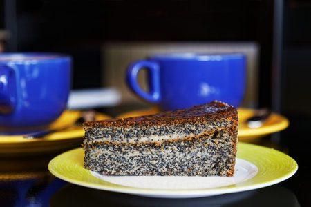 Closeup piece of poppy cake on a saucer opposite cups of coffee or tea on a dark blurry backgroundの写真素材