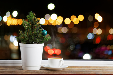 Small Christmas tree in a flowerpot decorated with one Christmas ball on a wooden window sill. The is is a cup of coffee. Outside the window are colored lights on the blurred background.の写真素材