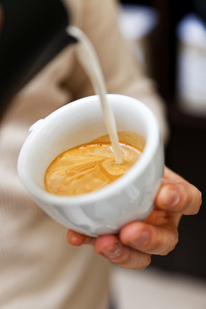 Closeup cup of cappuccino or latte in coffee-shop. Barista holding and pouring milk to make latte art. Shallow focus.の写真素材