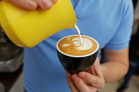 Closeup cup of cappuccino in coffee-shop. Barista holding and pouring milk to make latte art. Shallow focus.の写真素材