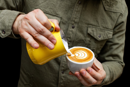 Closeup cup of cappuccino in hand of barista who pouring milk to make latte art. Shallow focus. Black background.の写真素材