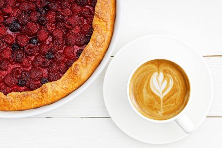 Closeup cup of coffee cappuccino and homemade galette with red raspberries and black currants on white wooden table. Top view.の写真素材