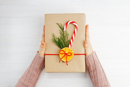 Female hands holding gift box wrapped in kraft paper, tied with red twine and decorated with a...