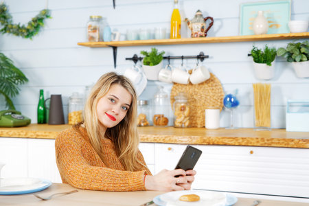 Young pretty girl sitting at a table in the kitchen with a phone in her handsの写真素材