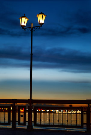 Vintage street lantern with warm yellow light and dark blue night cloudy sky on background. Night embankment river.の写真素材