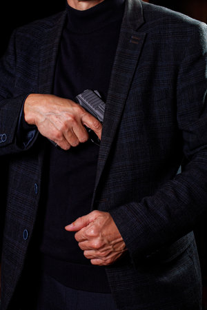 A man in a suit takes out a pistol from a jacket in darkness.