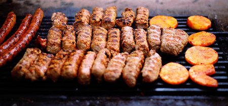 A close-up shot of grilled sausages and kebabs with sliced potatoes cooking on a charcoal grill.