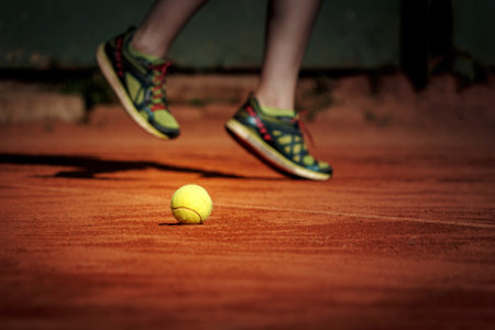 A tennis player jumps over a yellow tennis ball on a clay court.の写真素材