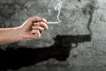 Male hand holding a cigarette, shadow on the wall with a gun, close-up, light background. Creative background. The concept of smoking kills, nekatin is poison, nicotine addiction. Copy space.の写真素材