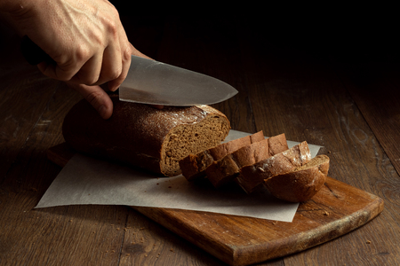 Creative background, male hands cut rye bread close-up, on a wooden cutting board, flat lay, copy space. The concept of fresh pastry, black bread.の写真素材