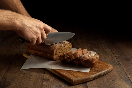 Creative background, male hands cut rye bread close-up, on a wooden cutting board, flat lay, copy space. The concept of fresh pastry, black bread.の写真素材