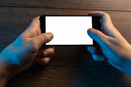 Closeup male hands holding smartphone with white screen, against the background of a wooden table. Concept of blank, mobile games, mockup, layout, copy space.の写真素材