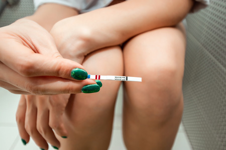 The girl makes a pregnancy test, two strips on the test. Waiting for the result, looking at the test strip. Close-up hands and legs. Concept of pregnancy, test, not desired baby.の写真素材