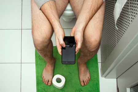 A man sitting on the toilet holding a smartphone in his hands, top view. The concept of problems with the chair, intestines, personal hygiene, constipation, boredom.の写真素材