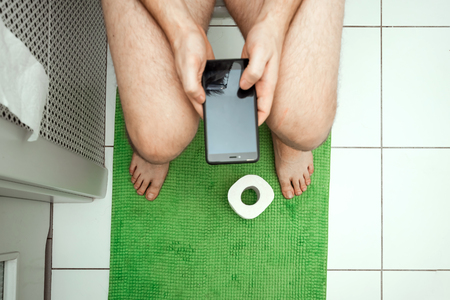 A man sitting on the toilet holding a smartphone in his hands, top view. The concept of problems with the chair, intestines, personal hygiene, constipation, boredom.の写真素材