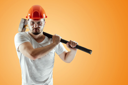 Bearded Man in a helmet holds a sledgehammer on an orange background. Concept of construction, contractor, repair.の写真素材