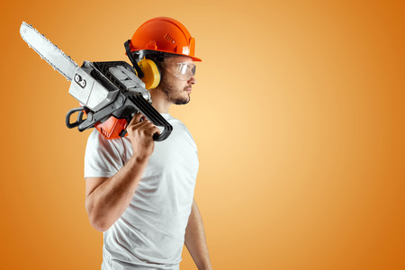 Bearded man in a helmet holds a chainsaw on an orange background. Construction concept, contractor, repair, lumberjack.の写真素材