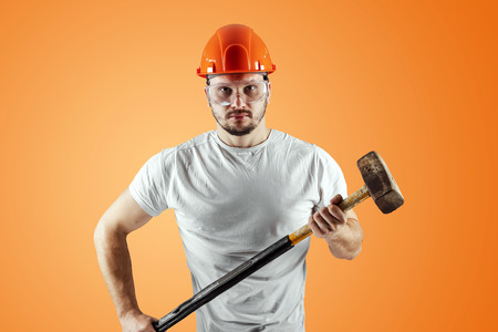 Bearded Man in a helmet holds a sledgehammer on an orange background. Concept of construction, contractor, repair.の写真素材