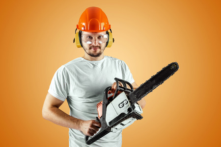 Bearded man in a helmet holds a chainsaw on an orange background. Construction concept, contractor, repair, lumberjack.の写真素材