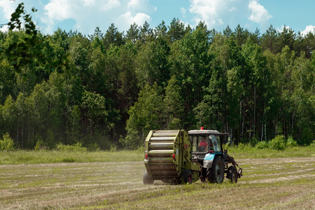 Agricultural machinery, a tractor collecting grass in a field against a blue sky. Hay harvesting, grass harvesting. Season harvesting, grass, agricultural land.の写真素材