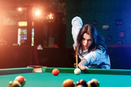 Young beautiful girl is playing billiards. Dark background. Pleasant pastime, rest, entertainment.の写真素材