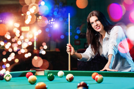Young beautiful girl is playing billiards. Dark background. Pleasant pastime, rest, entertainment.