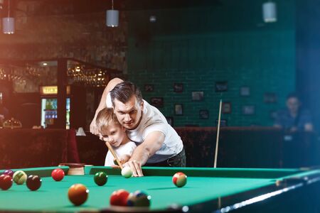 Father and son play billiards. The father teaches his son to play billiards. The concept of parents and children, upbringing, family rest.の写真素材
