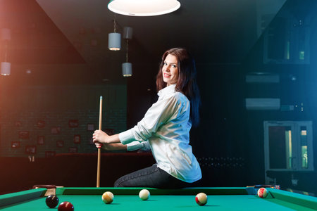 Young beautiful girl posing at a billiard table. Dark background. Pleasant pastime, rest,...